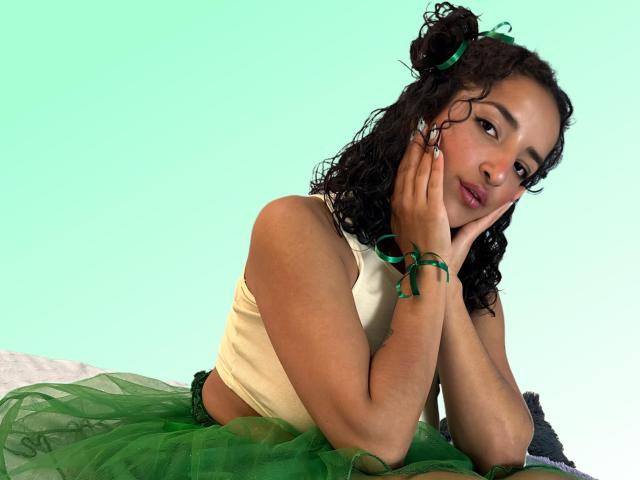 LunnaBenson with curly black hair, wearing a green tutu and a beige tank top, resting her chin on her hands, green bracelet, looking at the camera, soft smile.