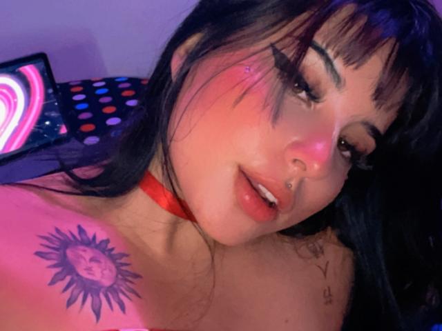 Grace Clarke, con capelli neri, guance rosate e un collare rosso, mostra un tatuaggio del sole sulla spalla, un piercing al naso e un bagliore rosa al neon, guarda la fotocamera con la bocca leggermente aperta..