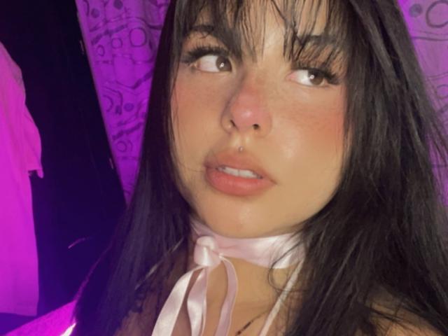 Grace Clarke tiene el cabello oscuro y lacio, un piercing en la nariz y lleva un collar de cinta blanca. Sus labios rosados están ligeramente entreabiertos, y tiene una mirada sutil y sensual..