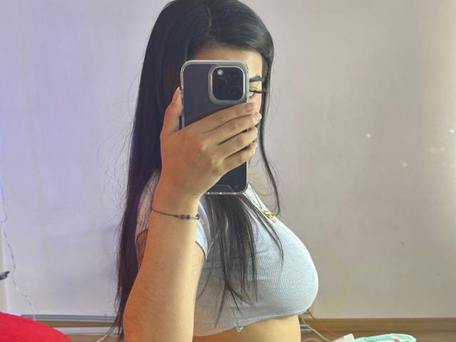 CamilePretty with long black hair, holding a phone, covering her face, wearing a tight gray top, showing her big boobs, in a mirror selfie.
