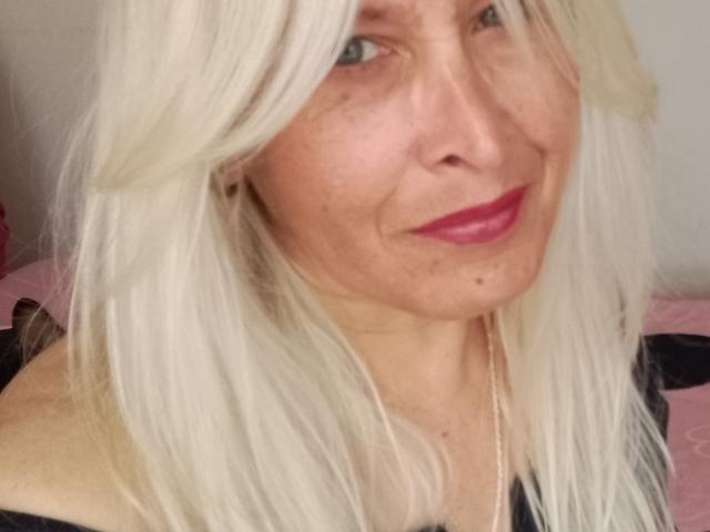 Rousk has white, straight hair, light skin, blue eyes, and pink lipstick. She's smiling with a small mole on her right cheek. She's wearing a black top.
