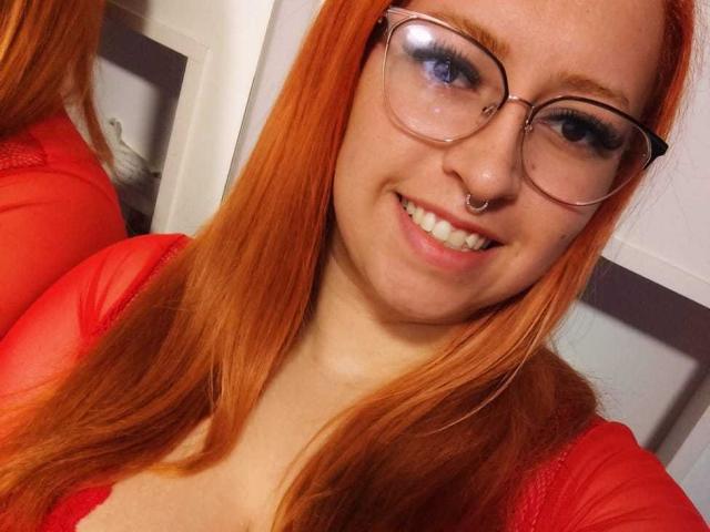 MelissaELuisa with red hair, glasses, nose ring, and a smile, wearing a red lace top, showing her tits in a selfie.