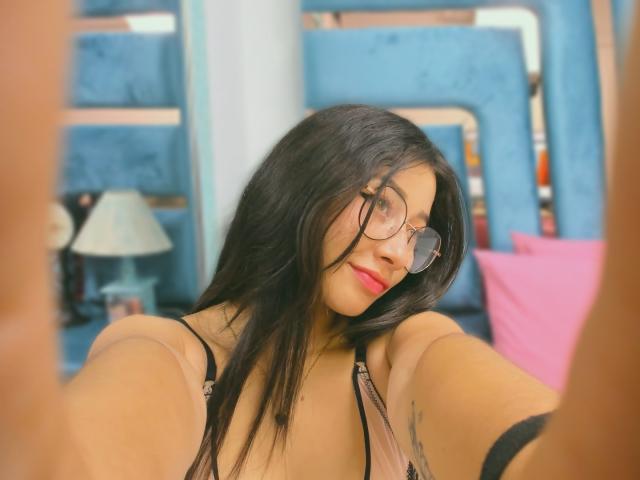 Genesiss with long black hair, glasses, and red lipstick, showing her tits in a beige top, taking a selfie, arms raised, blue headboard in the background.