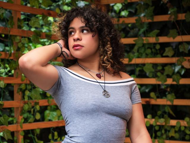 Curly-haired NeffellyRoss with medium tits, gray off-shoulder top, hand on head, looking to the side, wearing necklace, wooden lattice background, green leafy vines, pink lipstick, serious expression.