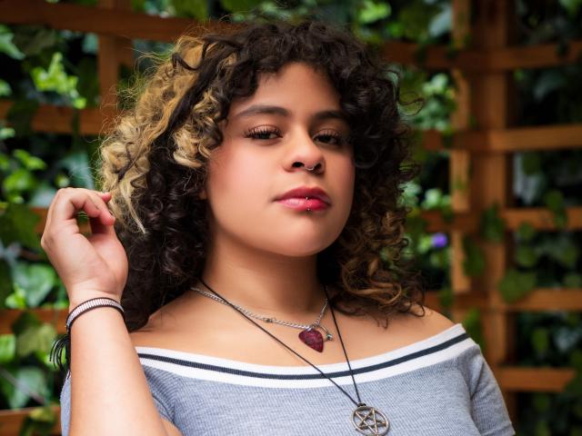 NeffellyRoss has curly brown and blonde hair, nose ring, pink lipstick, gray off-shoulder top, necklace with red pendant, and bracelet, standing with hand in hair, looking down.