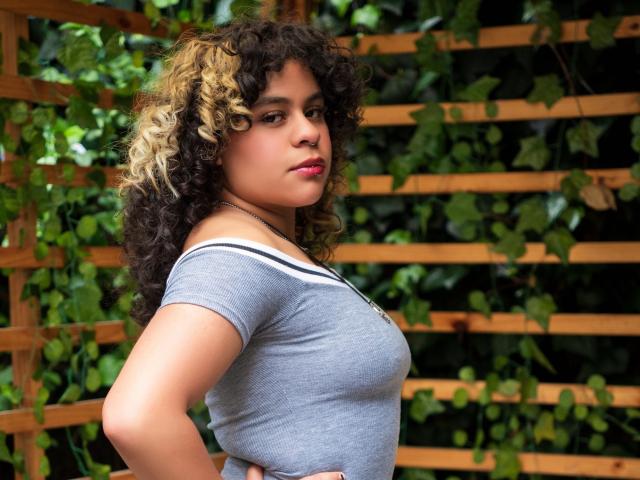 NeffellyRoss with curly brown and blonde hair, wearing a gray off-shoulder top, standing sideways, hands on hips, showing her tits and a confident, slightly smug expression.