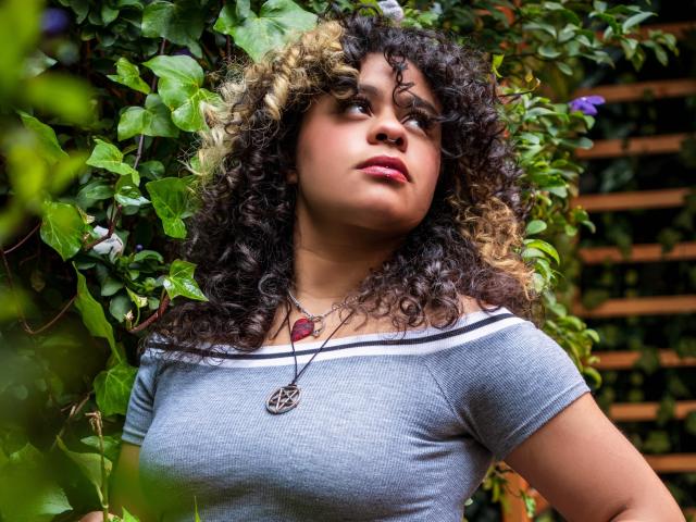 NeffellyRoss with curly brown and blonde hair, gray off-shoulder top, pink lipstick, necklace with red heart and pendant, green leafy background, looking up with a slight smile.