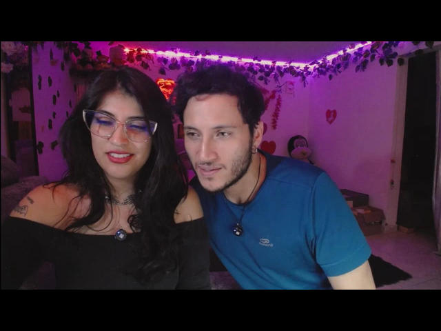 AzaharApolo con gli occhiali, una maglia nera a spalle scoperte e il seno in evidenza, e AzaharApolo con i capelli ricci, barba, una camicia blu e un piccolo pene, in una stanza illuminata di viola..