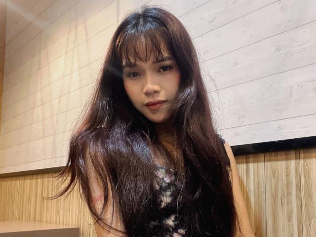FRESHotBHEAx-hot with long, dark, wet-looking hair, bangs, and small tits, wearing a black lace top, looking at the camera with a slight pout, in a wooden room.