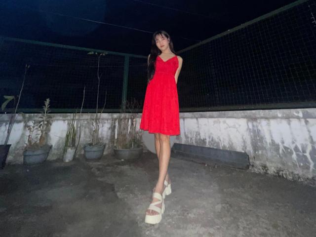 FRESHotBHEAx-hot in a red dress, standing with arms behind her back, wearing white platform sandals, on a dark rooftop with potted plants.