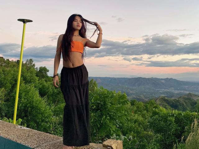 FRESHotBHEAx-hot with long dark hair, orange crop top, and black flowy pants, standing by a yellow pole, holding her hair, with a mountain view and sunset in the background.