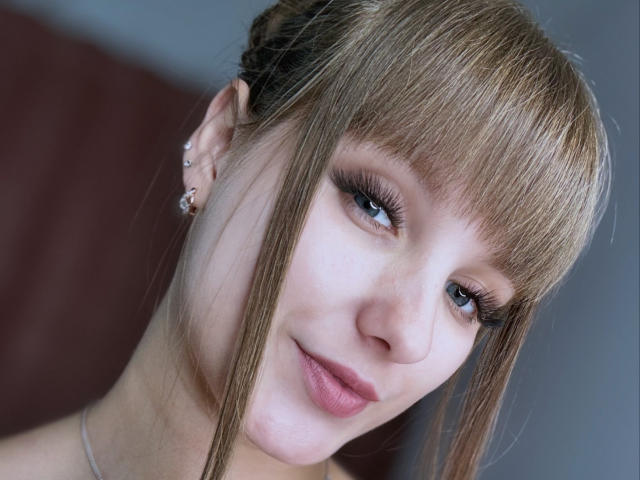 LeonaLoves has blonde bangs, blue eyes, and long eyelashes, with a subtle smile and light pink lipstick. She's wearing small earrings and a silver necklace.