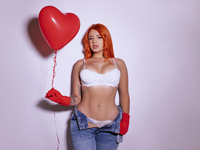 SherylRobhinss with red hair, white lace bra, and red gloves, holding a red heart balloon, unbuttoned jeans, showing her tits and white lace panties, standing against white.