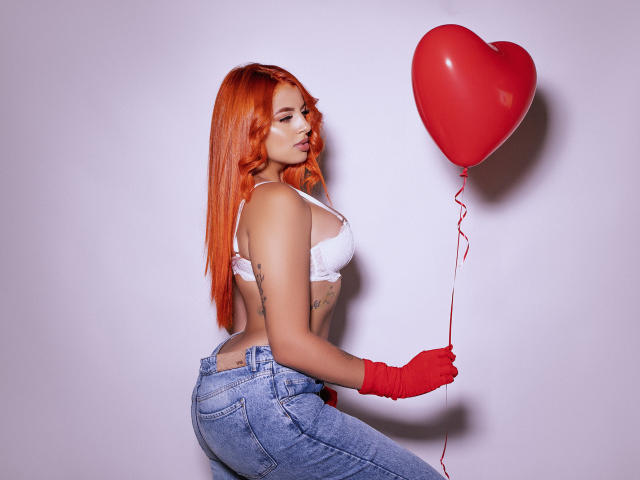 SherylRobhinss with orange hair, white bra, red gloves, and blue jeans, holding a red heart balloon, showing her ass and tattoos. Her tits are small, her pussy is unseen.