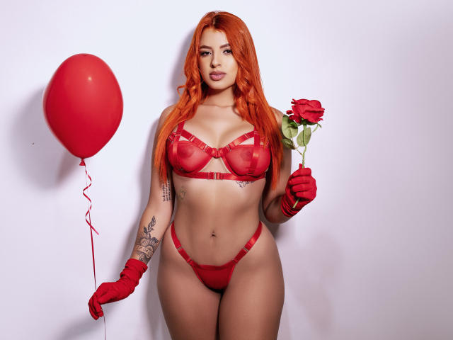 SherylRobhinss with red lingerie, red gloves, and red balloon, holding a rose. She has long red hair, tattoos on her right arm, and is posing confidently.