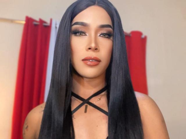 SeductiveKelly has long black hair, full eyebrows, heavy eyeliner, and glossy pink lips. She's wearing a black strappy top, and a small tattoo is visible on her chest.