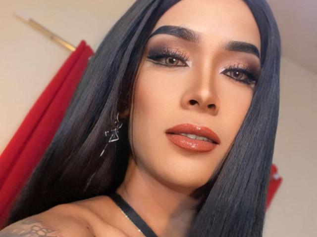 SeductiveKelly has long black hair, brown eyes with heavy eyeliner, and glossy orange lipstick. She's wearing black earrings and has a tattoo on her visible shoulder.