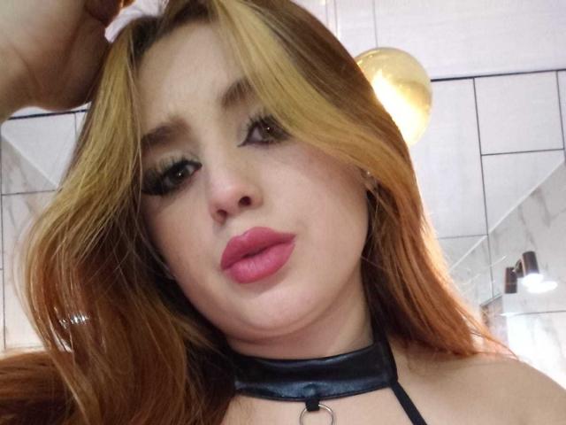 StephanieMoretti with blonde hair, pink lips, black leather choker, and long eyelashes, looking at camera, hand in hair, light skin, and a slight pout, in a bathroom.