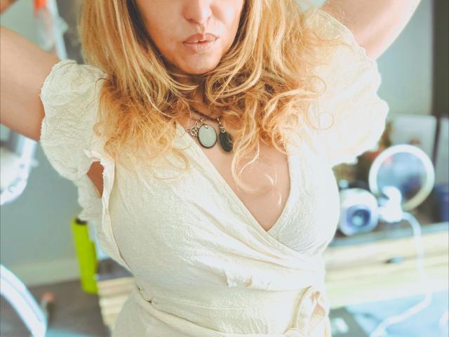 ZoeRubia-hot with blonde curly hair, wearing a white dress with a deep V-neck, showing her large tits, arms raised, necklace with a circular pendant, pouty lips.