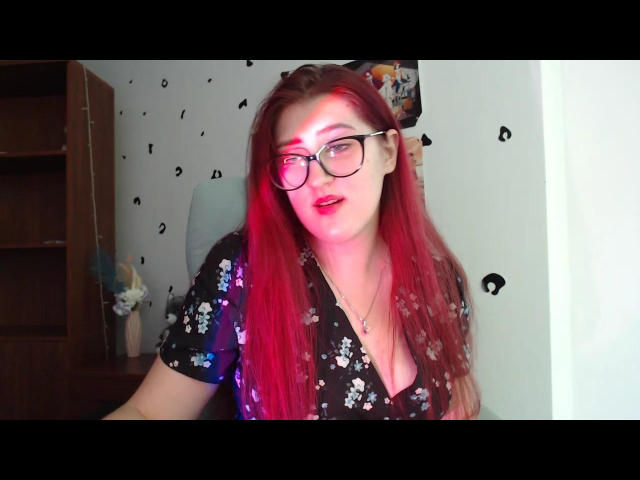 Triziya with long red hair, glasses, and a black floral shirt, showing off her tits in a selfie. Red light on her face, slightly open mouth, casual expression.