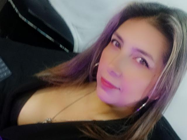 Michelle Dumont tem cabelo castanho, pele rosada e está vestindo uma blusa preta com zíper, que mostra um pouco do colo. Ela tem batom rosa e está sorrindo levemente..