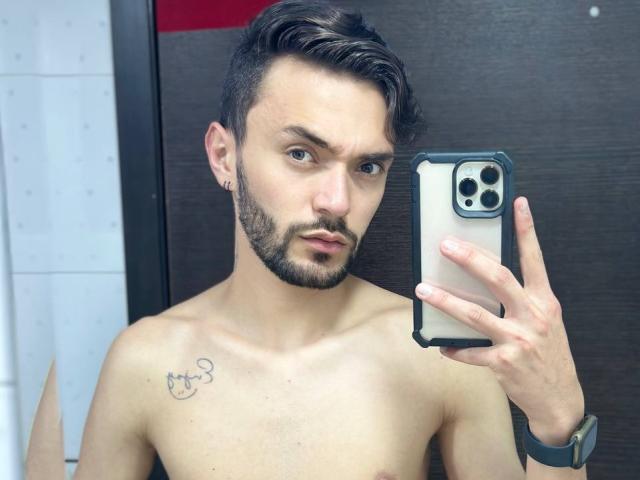 Shirtless JakeMagnus with short dark hair, beard, tattoo of a cat on left chest, holding phone, wearing watch, looking at mirror, taking selfie. Boobs: none, Ass: none, Tits: none.