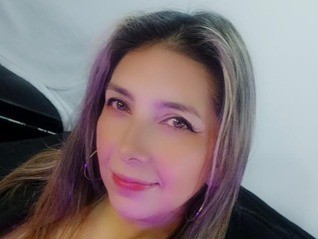 Michelle Dumont tem cabelos castanhos longos com mechas loiras, usa brincos de argola roxos e está sorrindo com batom rosa. Seus olhos castanhos são ligeiramente inclinados para cima, e ela tem um rosto bonito..