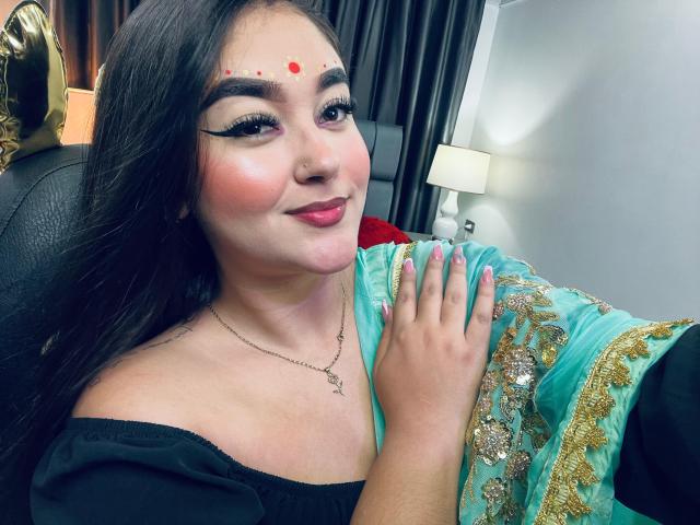 AnaShalimarr with long black hair, red dot on forehead, black off-shoulder top, green and gold embroidered dress, pink blush, smiling, hand on chest, eyeliner, pink lipstick.