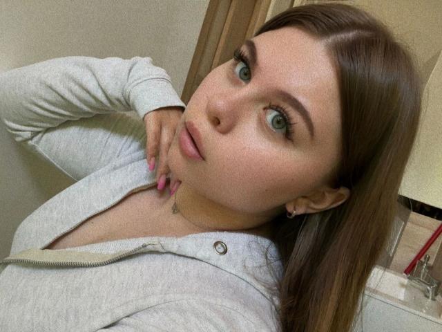 AbbyKuss-hot with green eyes, pink nails, and a gray zip-up hoodie, touching her neck with a hand, showing a small tattoo and earrings, in a bathroom selfie.