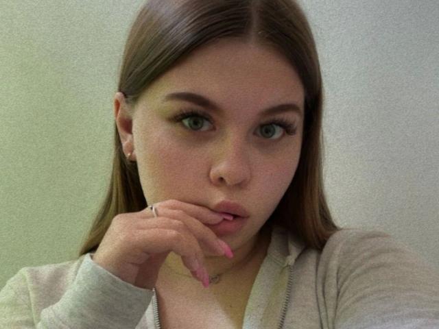 AbbyKuss-hot with long brown hair, green eyes, and pink nails, biting her finger, wearing a grey hoodie, looking at the camera with a slightly parted pink mouth.