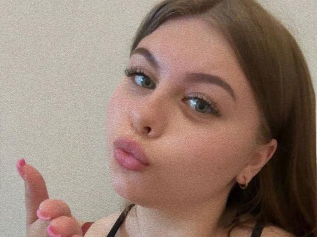 AbbyKuss-hot with pouty lips, brown hair, green eyes, and pink nails, giving a middle finger, has small earrings, and is wearing a black halter top.