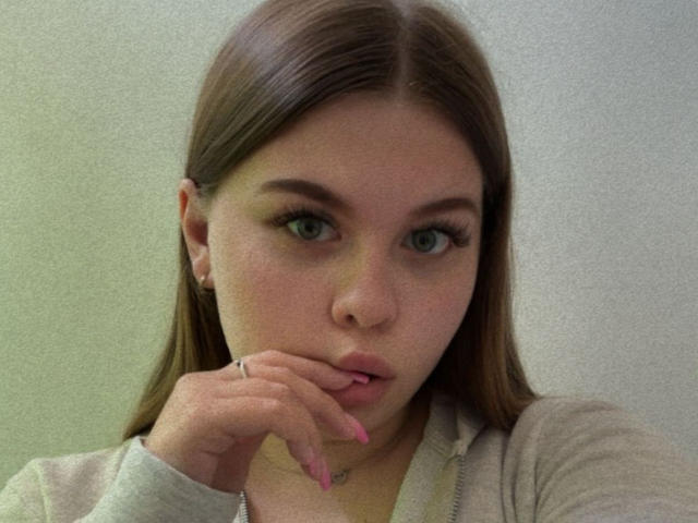 AbbyKuss-hot with long brown hair, green eyes, and pink lipstick, biting her finger with a ring on her hand, wearing a gray hoodie, looking at the camera.