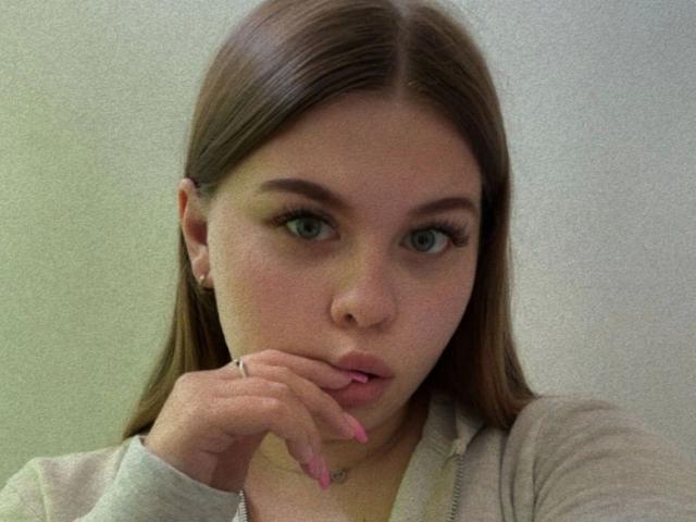 AbbyKuss - aantrekkelijk, met bruin haar, groene ogen en roze nagels, draagt een grijze hoodie, raakt haar lip aan met haar vinger en kijkt naar de camera..