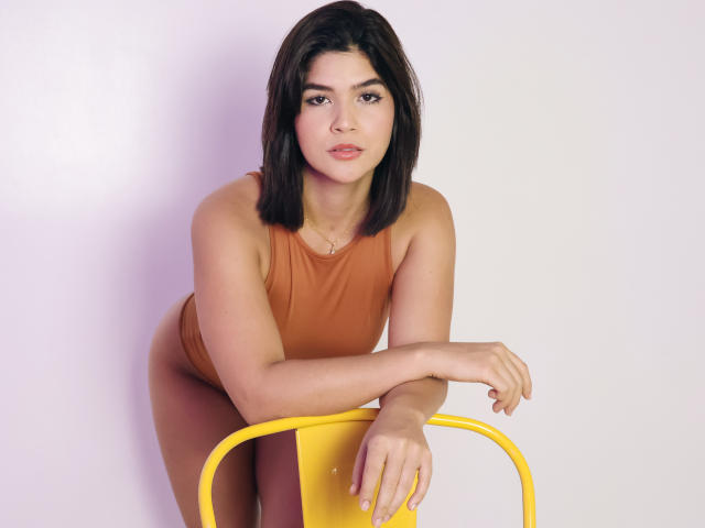 ScarlletZhen with shoulder-length black hair, brown tank top, and yellow chair, leaning forward, hands on chair, looking at camera with a slight smile, soft pink lips.