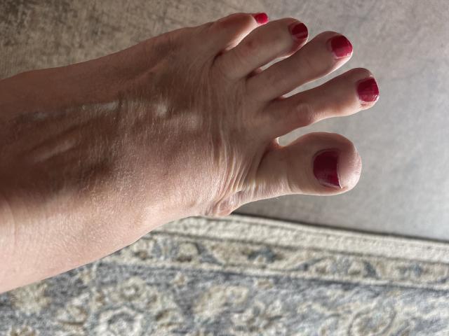 Bourgeoisex's foot with red-painted toenails, showing detailed skin texture and slight wrinkles, positioned on a carpeted surface.