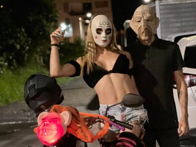 MyaAndJack69 in a black bra and denim shorts, holding a cigarette, with a white mask and red cheeks, stands next to an old dude in a black shirt.
