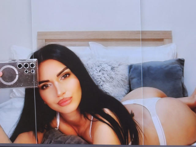 Paola Adams, con lunghi capelli neri, sdraiata su un letto, indossa biancheria intima bianca, mostra il sedere, tiene in mano un telefono con un design a forma di anello, e guarda la fotocamera..