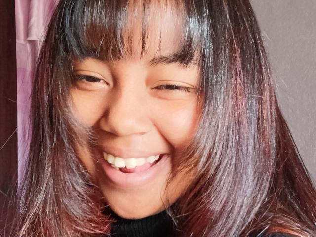 MaliciaQueen's smiling face with straight, dark brown hair, slightly wavy ends, and a black turtleneck, winking with her teeth showing. Her skin is warm brown.