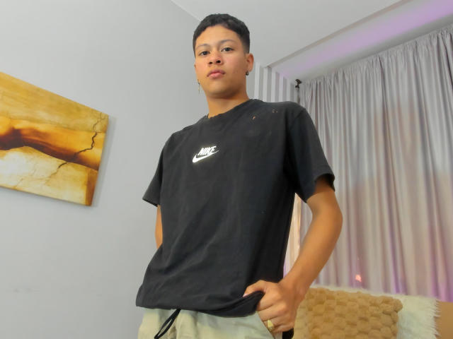 Jonas Escalante, capelli neri corti, indossa una maglietta nera Nike, pantaloni verdi, in piedi con le mani sui fianchi, guarda la telecamera, tenda bianca sullo sfondo, cuscino beige a destra..