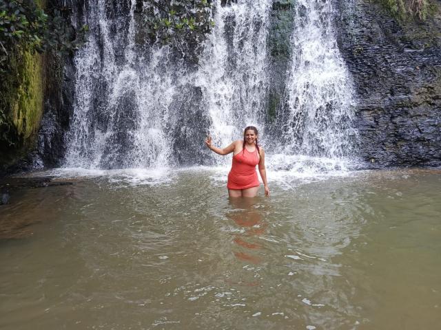 DylaraCollins stands waist-deep in a waterfall, wearing a red tank top, water dripping from her brown hair, wading with one hand raised, her ass partially submerged.