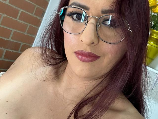 DylaraCollins with shiny red lipstick, dark makeup, and round glasses, has dark red hair and is topless, showing her bare shoulders and small, cute tits.
