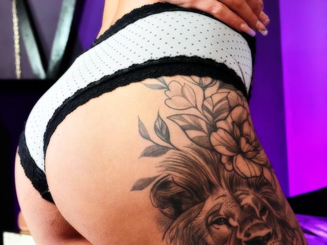 Las panties blancas con lunares negros de NenaBlack, con encaje negro, muestran un tatuaje detallado del rostro de un león y flores en su trasero bronceado y curvilíneo. Su mano descansa sobre su parte baja de la espalda..
