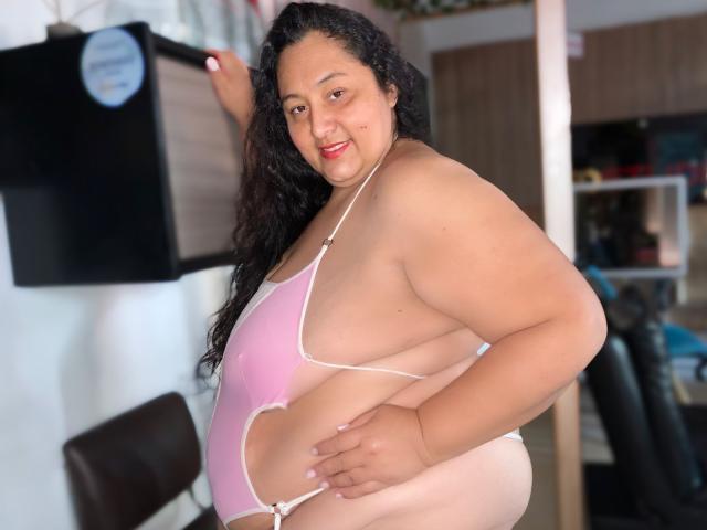 TistMature, a curvy woman with long black curly hair, wearing a pink and white, side-cut, high-cut swimsuit, showing her big ass and small tits, smiling at the camera.