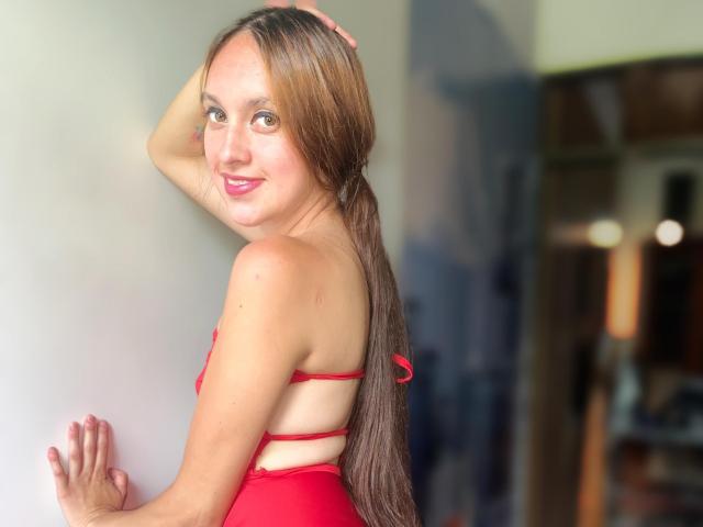 SofySweety with long brown ponytail, red strappy bikini, smiling, hand on wall, looking back, showing her tits, pink lipstick, light skin, brown eyes, indoors, soft light.
