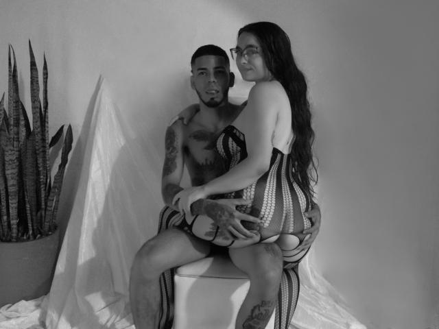 MiaAndLeon with long dark hair, glasses, and striped lingerie, sitting on Leon's lap, who's shirtless with tattoos, holding her butt. Potted snake plant on left. Black-and-white photo.