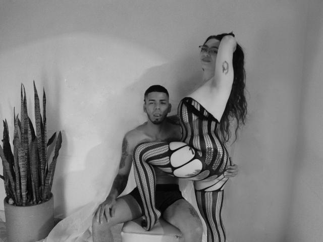 MiaAndLeon with striped bodysuit, raised arm, and glasses, sits on Leon's lap, who's shirtless in dark shorts, holding her up. Snake plant on left. Black-and-white photo.