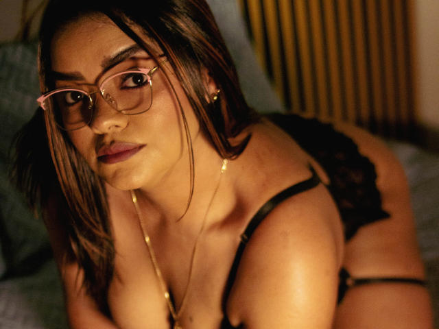 NinnaRose with brown hair, glasses, and gold necklace, wearing black lace lingerie, looking at camera with slightly parted lips, showing her tits and ass in dim lighting.