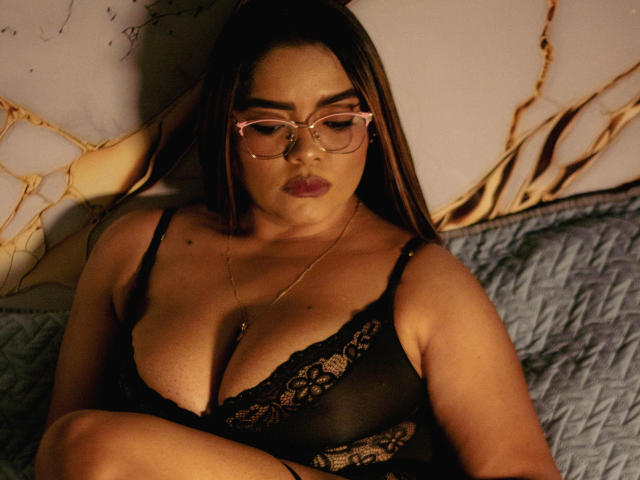 NinnaRose with large tits, wearing black lace lingerie and pink glasses, sitting on a quilted blue bed, looking down with a serious expression, long brown hair, gold necklace.