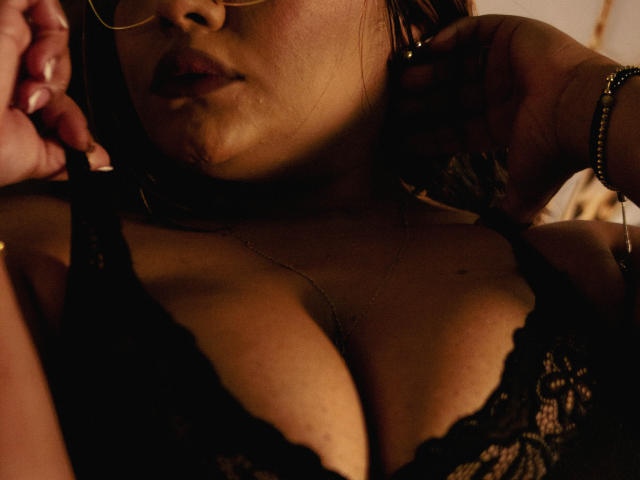 NinnaRose with her face partially in shadow, wearing glasses and black lace lingerie, showing her large tits, her hand near her ear with a bracelet on her wrist.