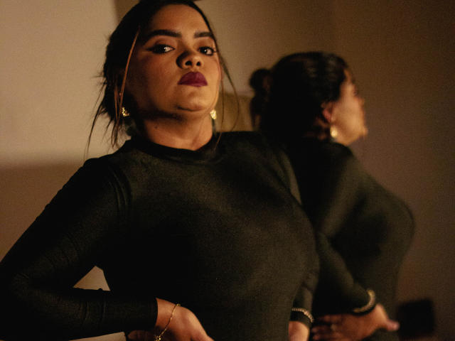 NinnaRose with dark brown skin, wearing a black long-sleeve top, standing with hands on hips, reflection in mirror, looking confident, red lipstick, gold earrings, dim lighting.